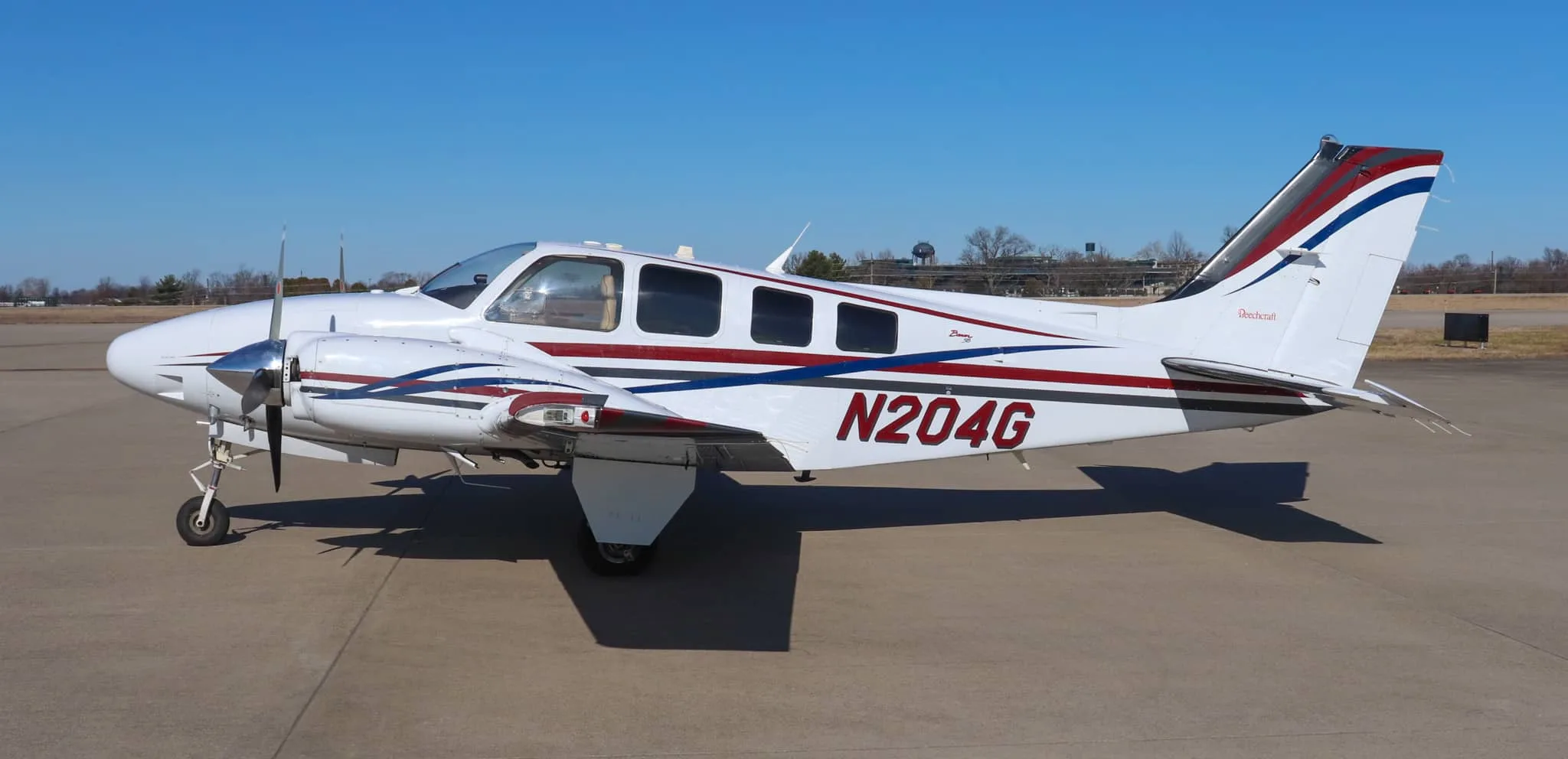 Side view of a white twin-engine Beechcraft Baron N204G with red, blue, and gray stripes, parked on an airfield.
