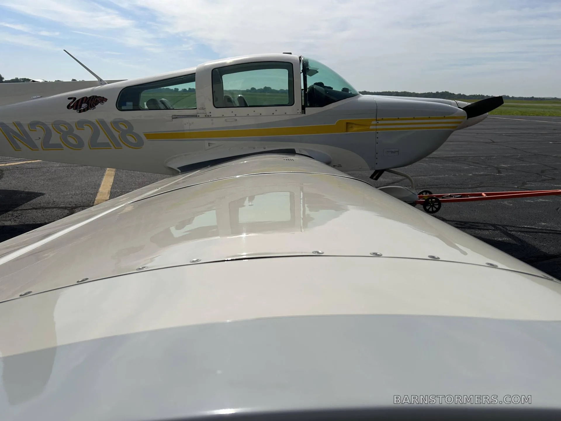 An overhead view of the wing and fuselage of a white and yellow Grumman AA-5B Tiger aircraft with tail number N28218 on an airfield.