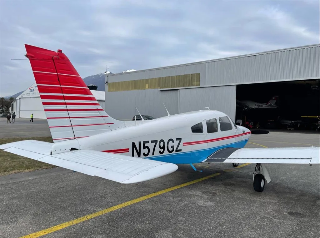 A white and blue Piper PA-28-181 Archer II with red stripes on the tail, parked on an airfield next to a hangar.