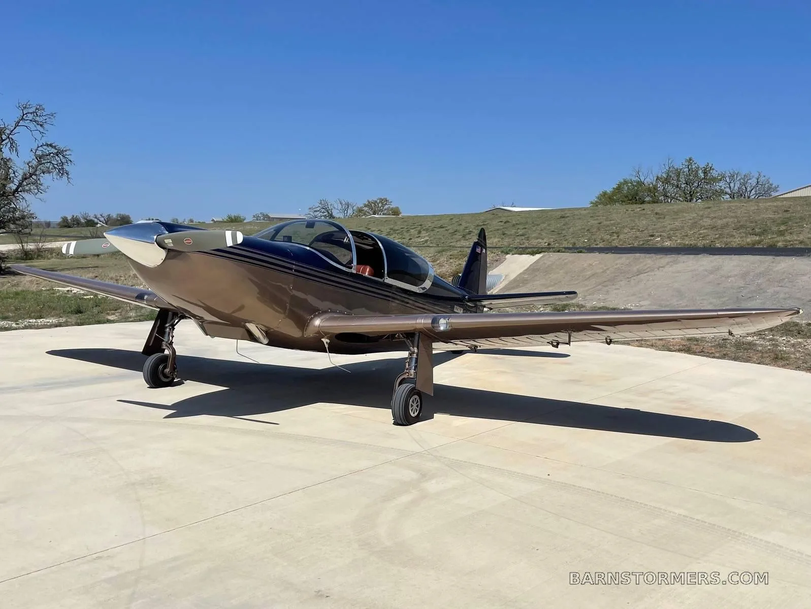 A bronze and dark blue single-engine aircraft with a polished nose cone parked on a concrete pad under a clear blue sky.