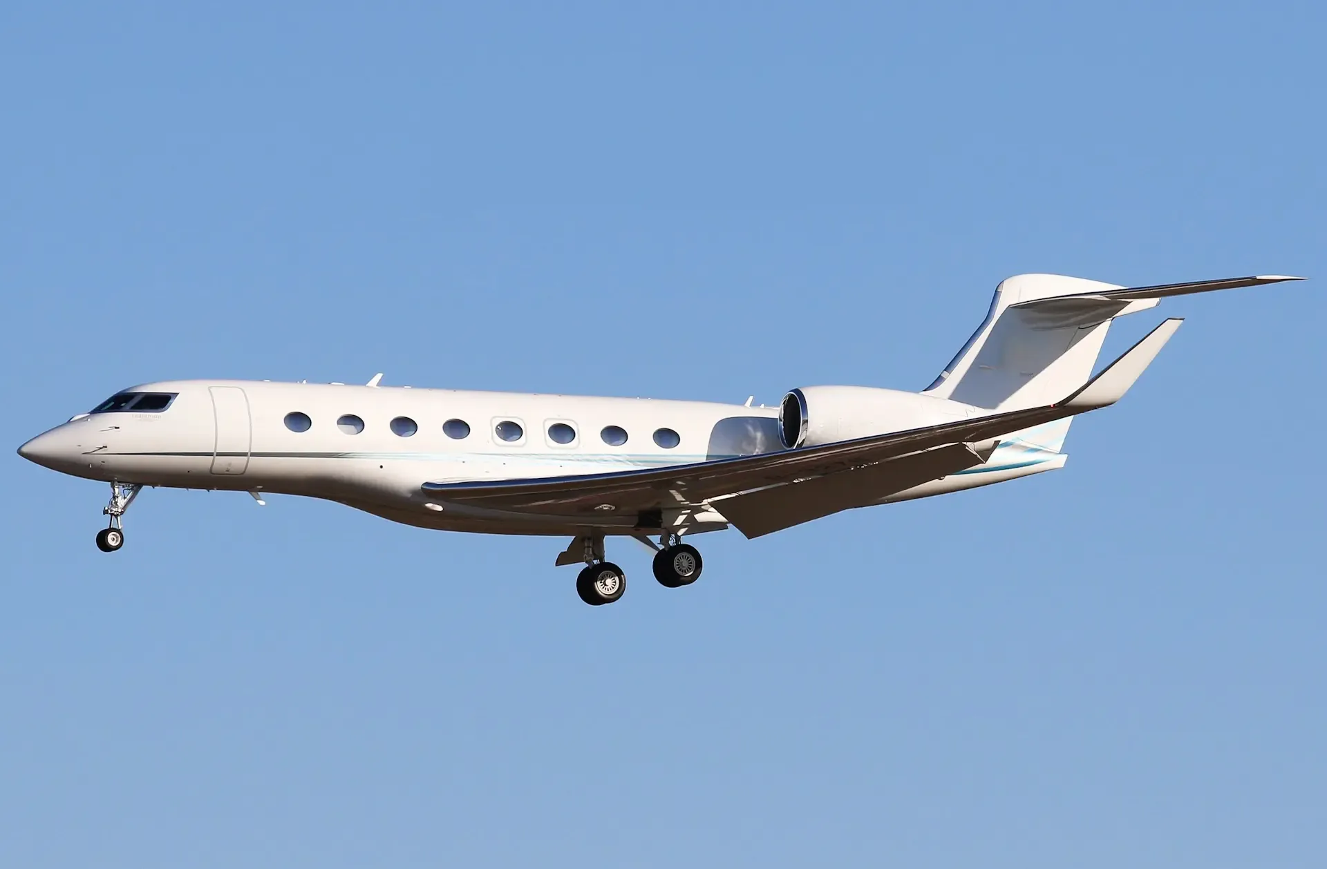 A white Gulfstream business jet with landing gear extended flies against a clear blue sky.