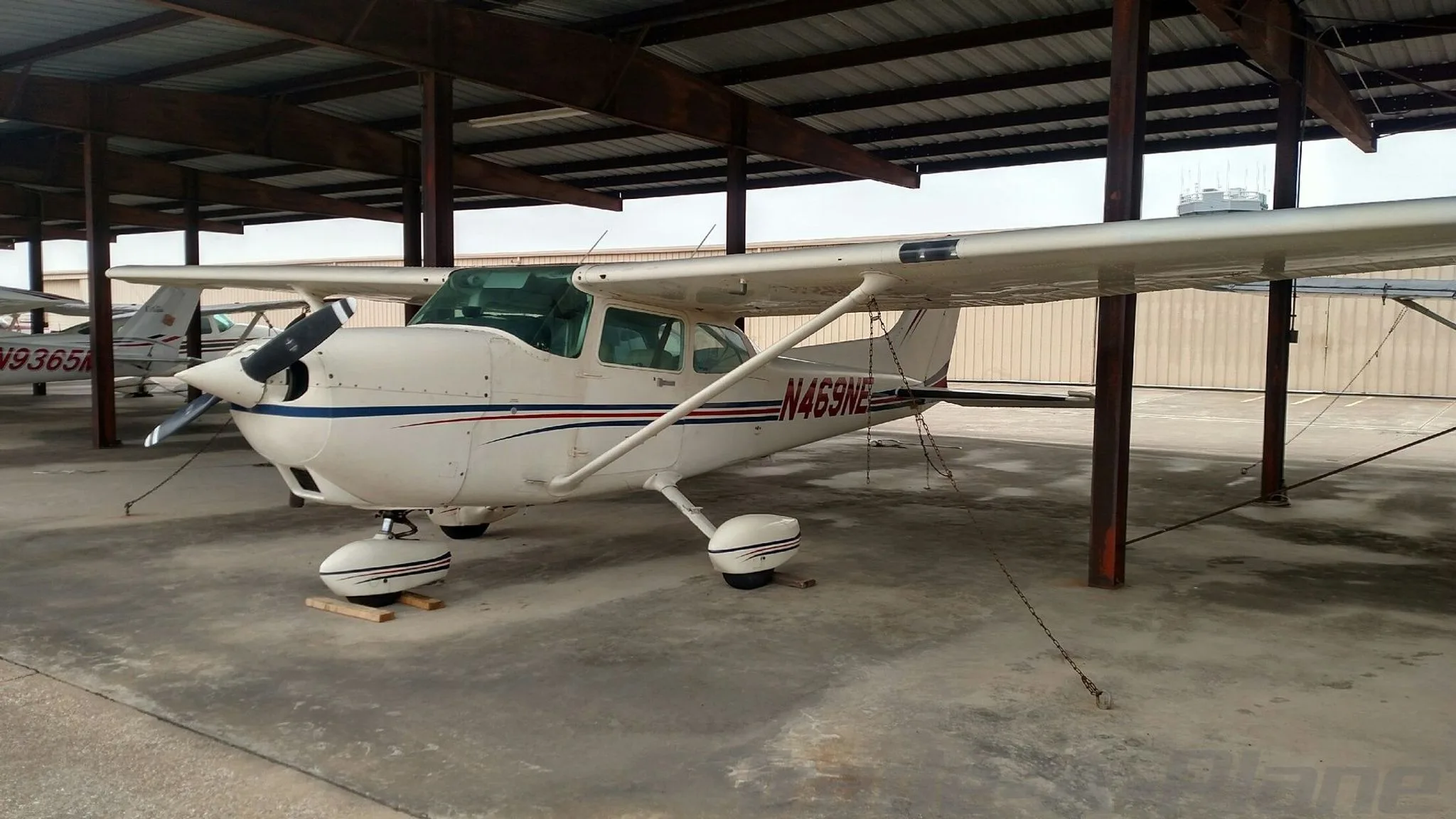 A white Cessna 172 with blue and red stripes parked under a metal hangar, showing the right side.