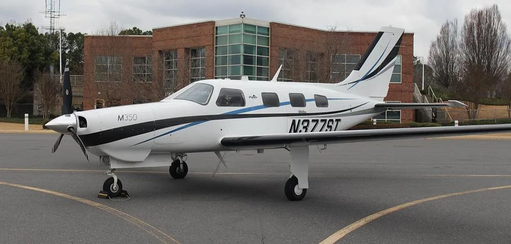 A white Piper M350 aircraft with black and blue stripes, registration N377ST, parked on an asphalt tarmac in front of a brick building.