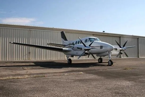 A white twin-engine turboprop aircraft with black and grey stripes, parked on an asphalt ramp in front of a metal hangar.