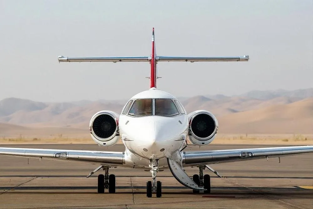 A head-on view of a white business jet with red and black stripes, parked on a tarmac with distant mountains.
