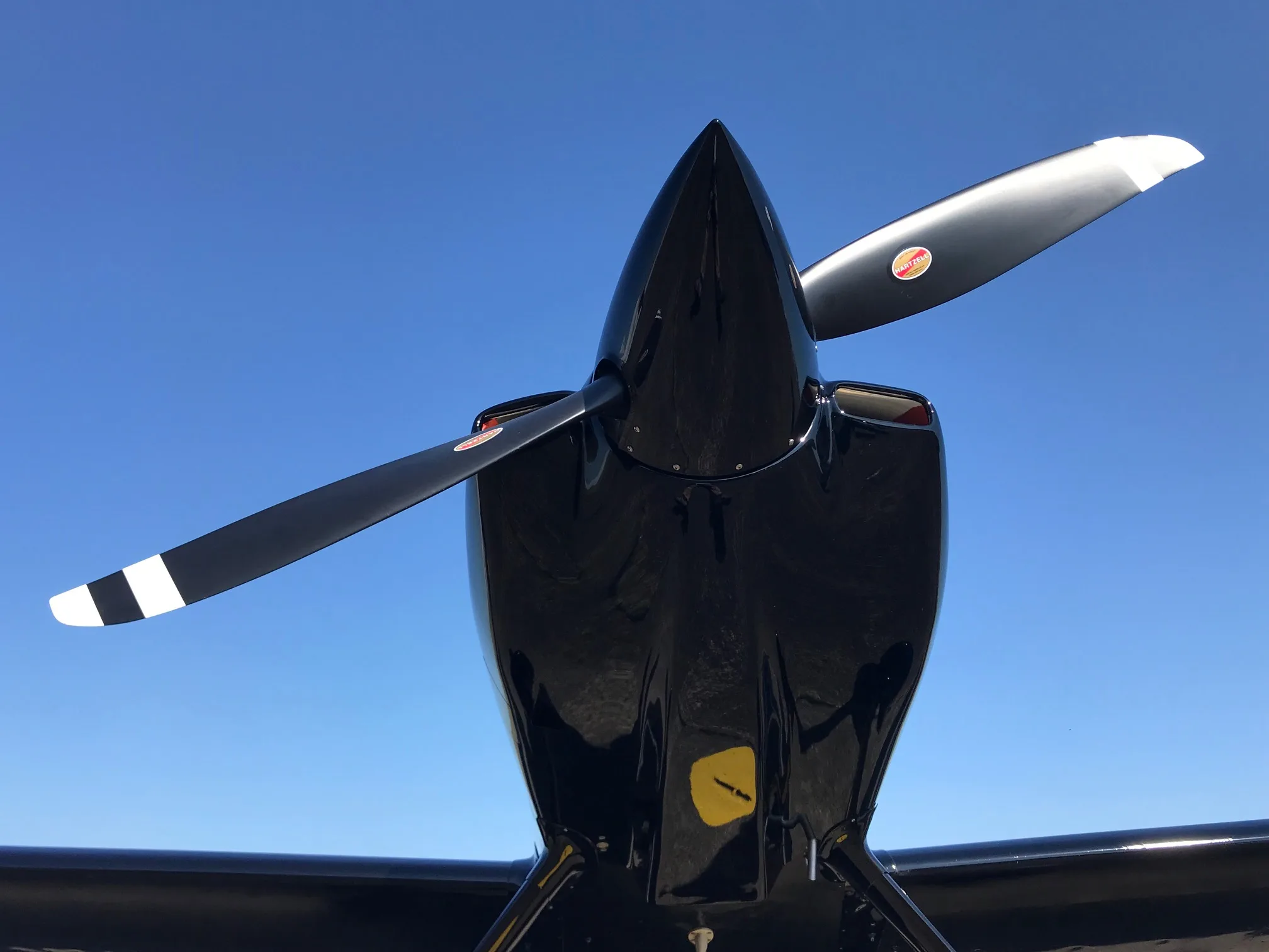 A front view looking up at the black experimental aircraft's nose and propeller against a clear blue sky.