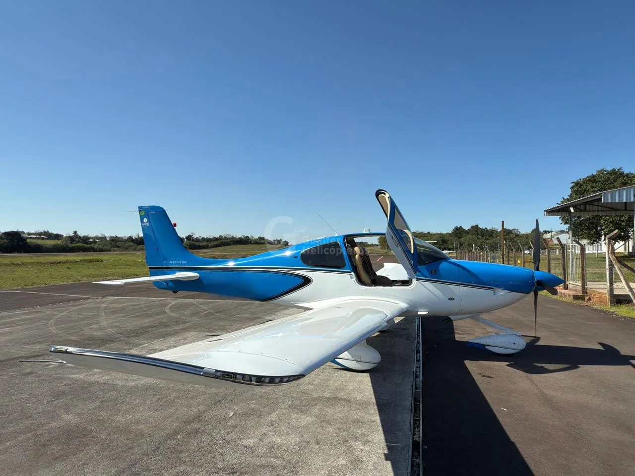 A blue and white Cirrus SR22 aircraft with its doors open, parked on an airfield tarmac under a clear blue sky.