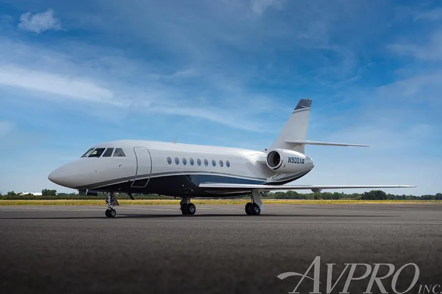 A Dassault Falcon business jet, registration N902AM, parked on an airfield under a blue sky, showing its white and dark blue paint scheme.