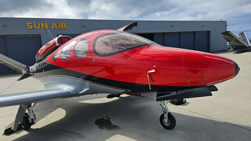 A close-up front view of a red, black, and silver Cirrus Vision Jet (N26VJ) parked in front of a "SUN AIR" hangar.