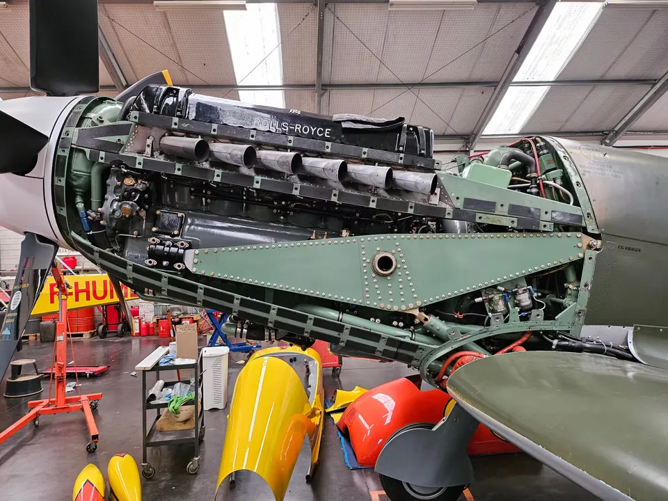 The exposed Rolls-Royce engine of a Supermarine Spitfire undergoing maintenance in a hangar, with cowling panels removed.