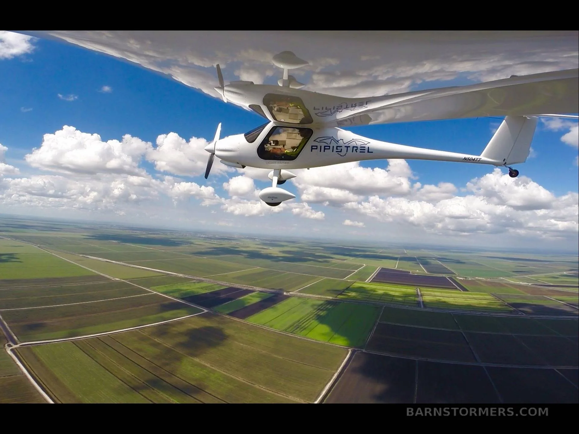 A white Pipistrel aircraft flying over green agricultural fields under a partly cloudy blue sky.