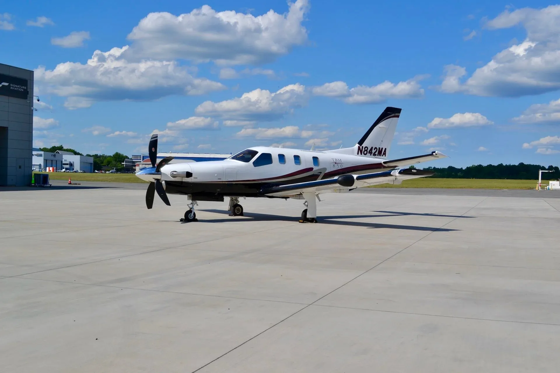 A white TBM 850 aircraft with black and maroon stripes is parked on an airport tarmac under a blue sky with clouds.