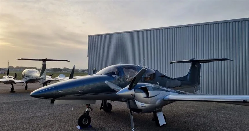A dark blue and silver twin-engine propeller aircraft is parked on an airfield next to a hangar at sunset.