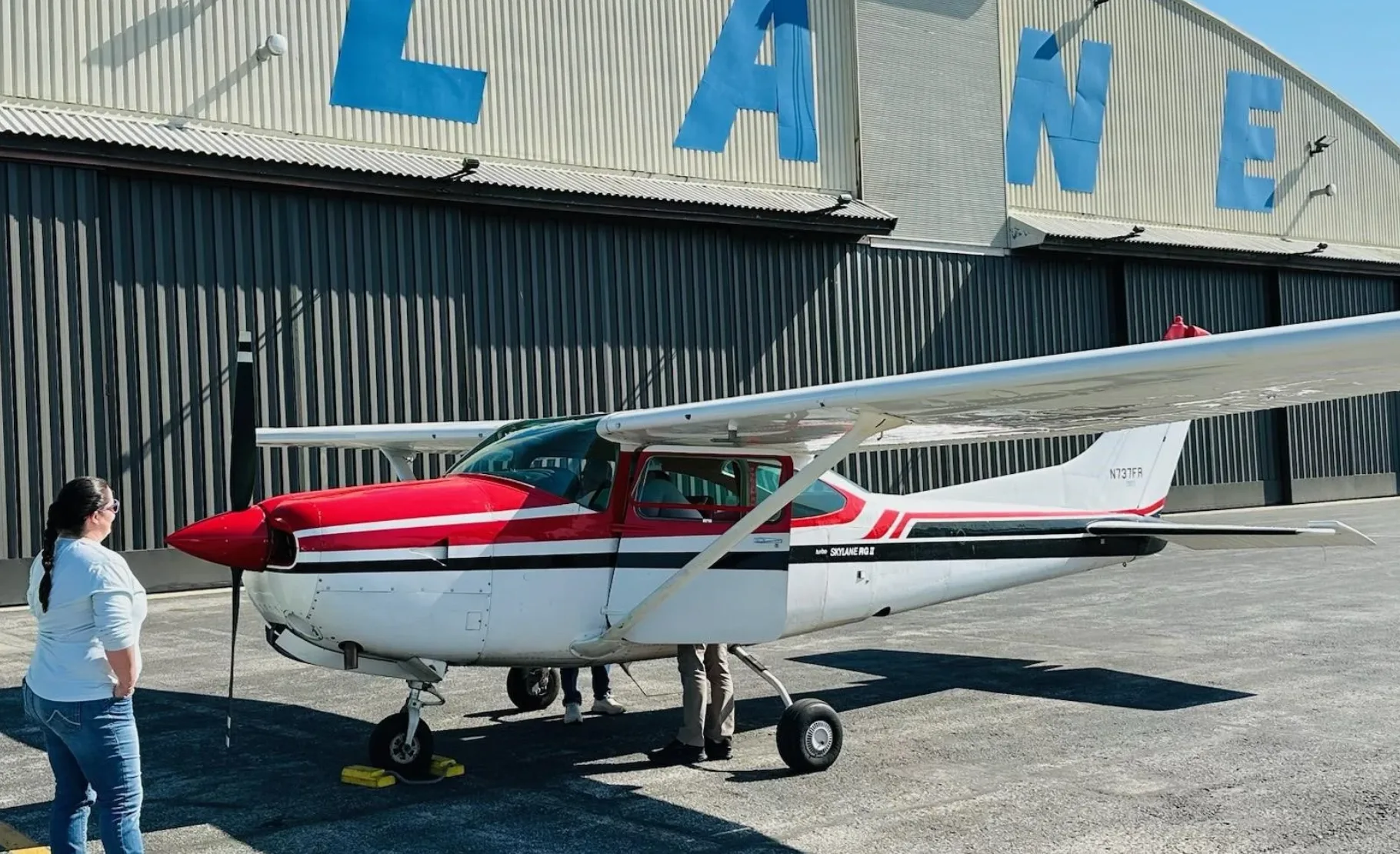 A red and white Cessna 182 Skylane (N737FR) parked outside a hangar with people nearby.