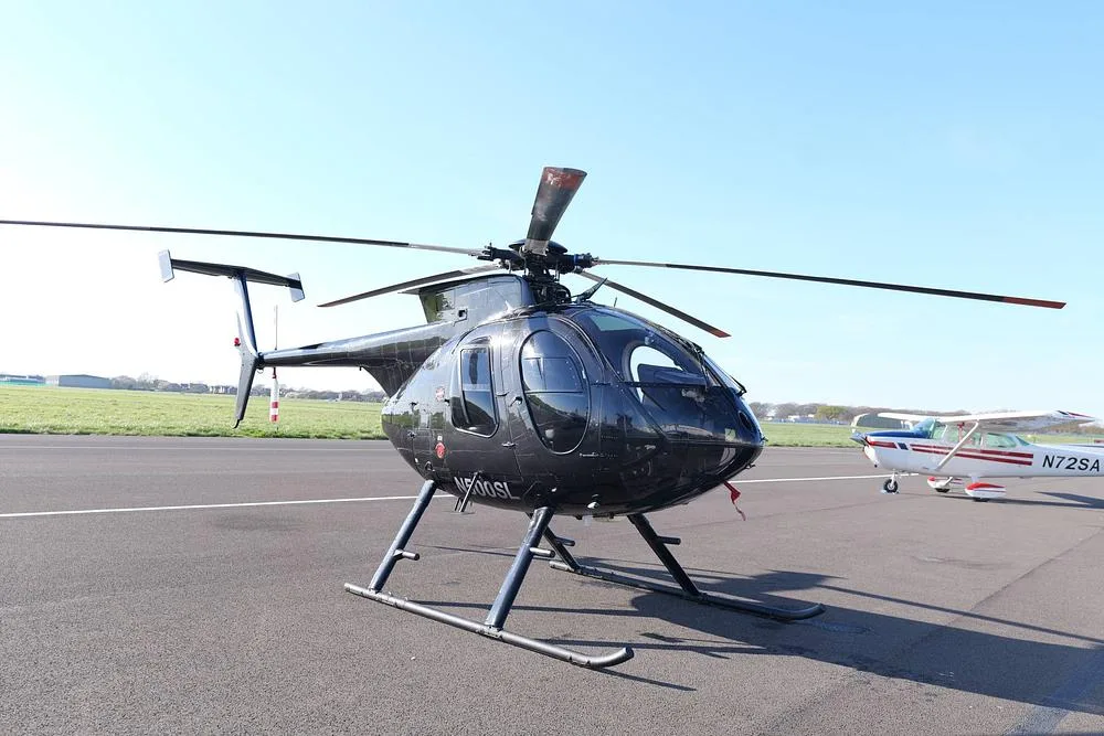 A black MD 500 helicopter (N500SL) is parked on an airfield tarmac with a small white plane in the background.