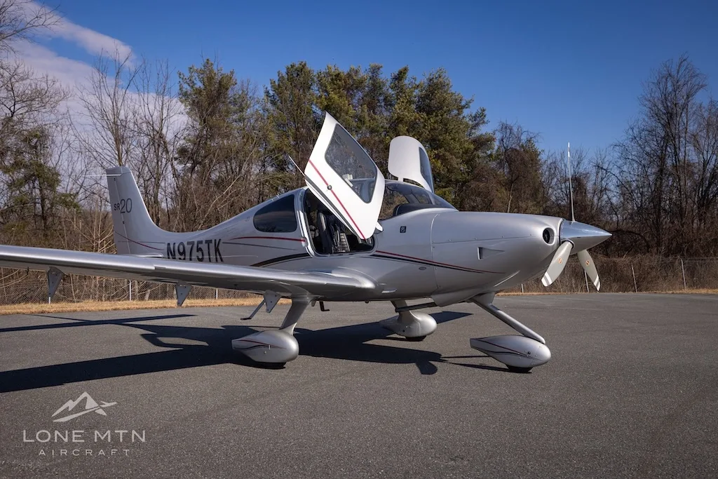 A silver Cirrus SR20 aircraft with registration N975TK parked on an asphalt runway, viewed from the front-right with doors open.