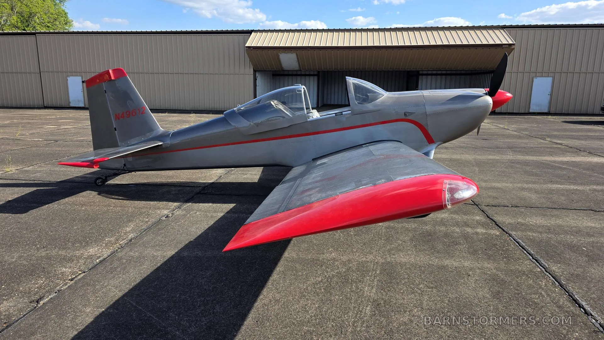 A side view of a silver single-engine aircraft with red wingtips and tail accents, parked on an asphalt tarmac next to a hangar.