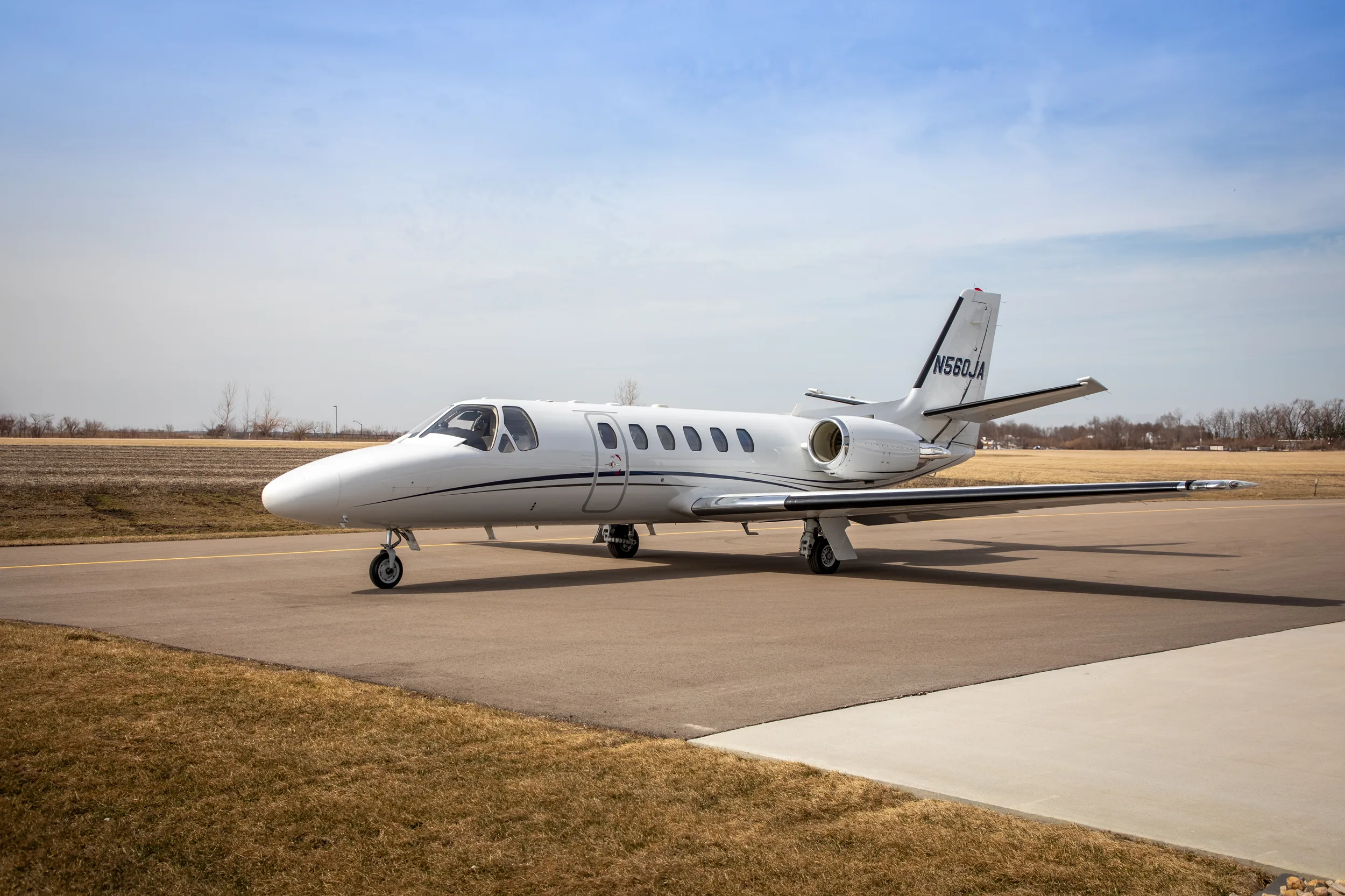 A white Cessna Citation Excel (N560JA) with black and grey stripes parked on an airfield with fields in the background.