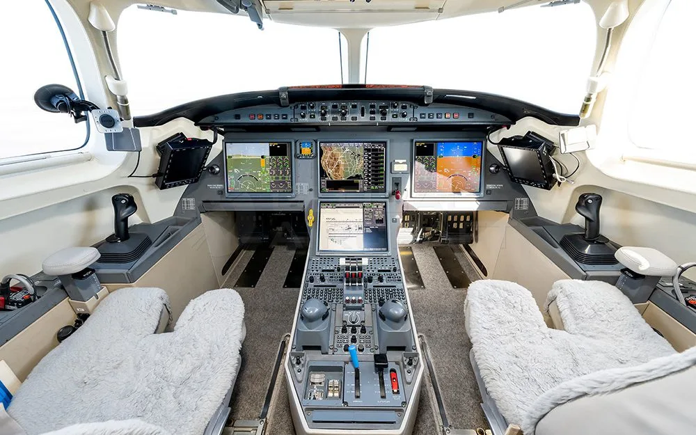 The modern cockpit of a private jet, featuring multiple digital displays and two pilot seats with sheepskin covers.