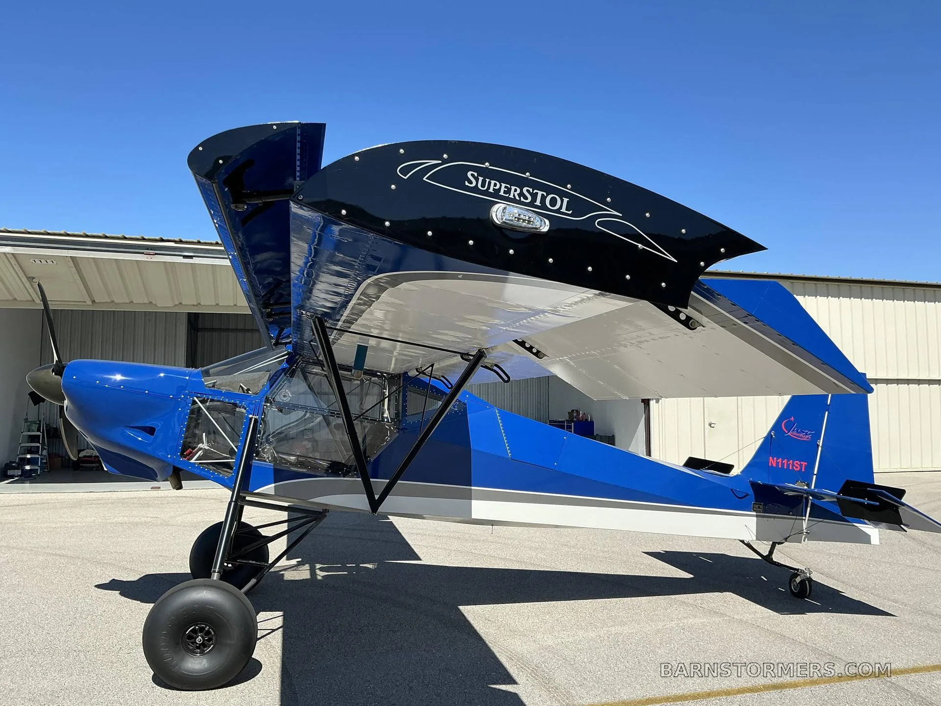 A blue and white SuperSTOL aircraft with its top wing panels open, parked on an asphalt apron in front of a hangar.