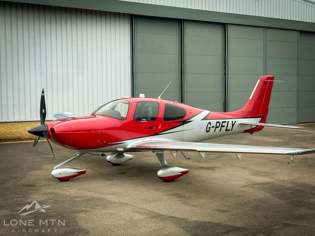 A red and white Cirrus SR22 aircraft (G-PFLY) parked on a tarmac in front of a hangar.