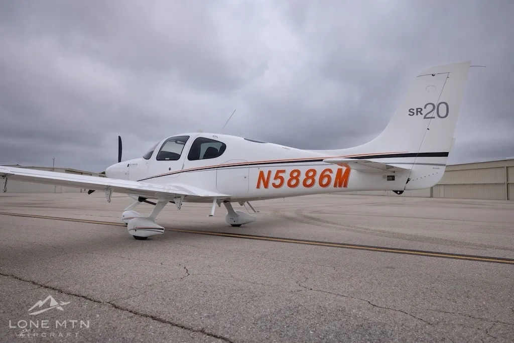 A side view of a white Cirrus SR20 aircraft with orange and black stripes, registration N5886M, on an airfield.