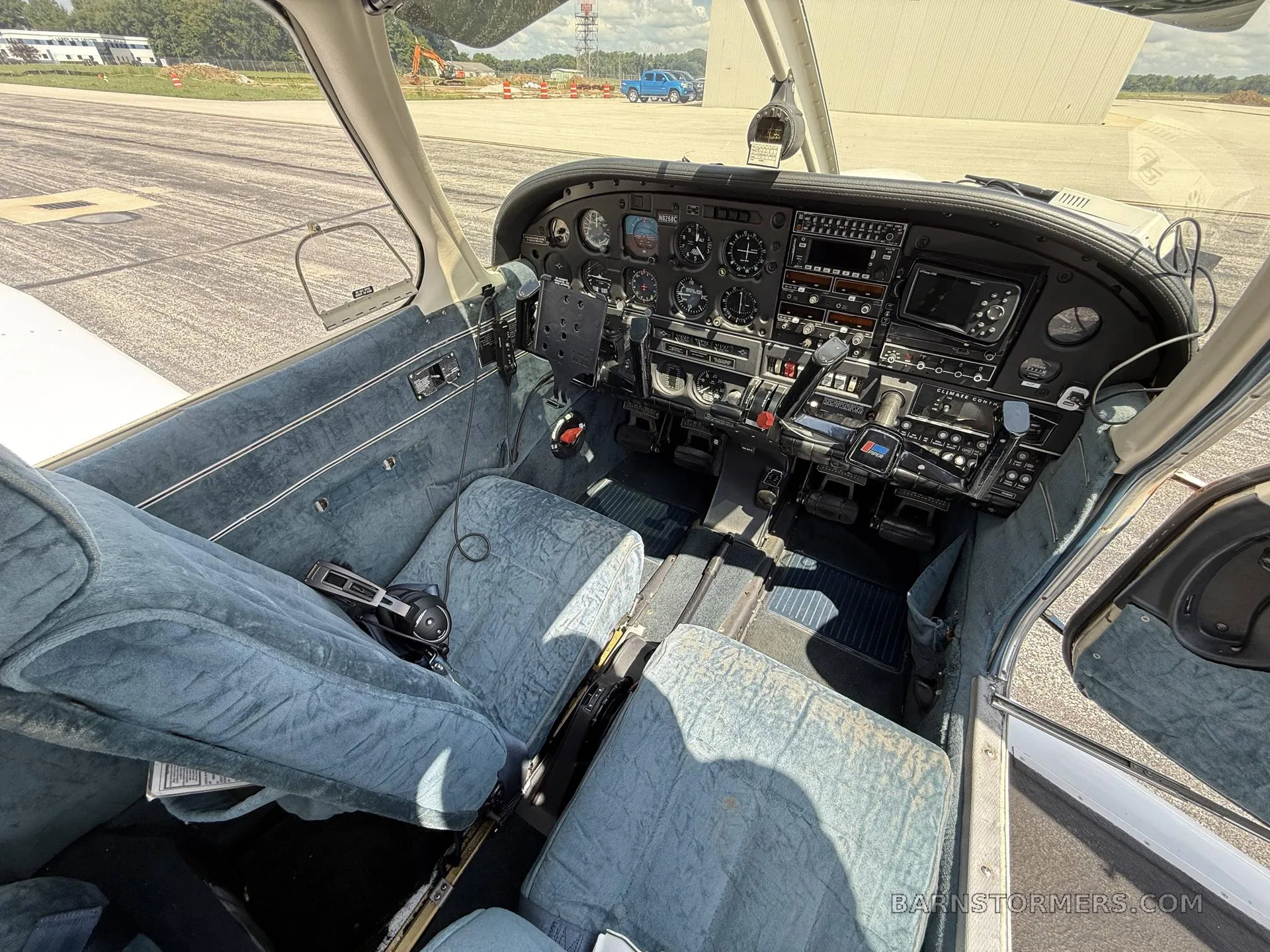 The interior of a Piper aircraft showing the cockpit and blue fabric front seats, with an airfield visible outside.