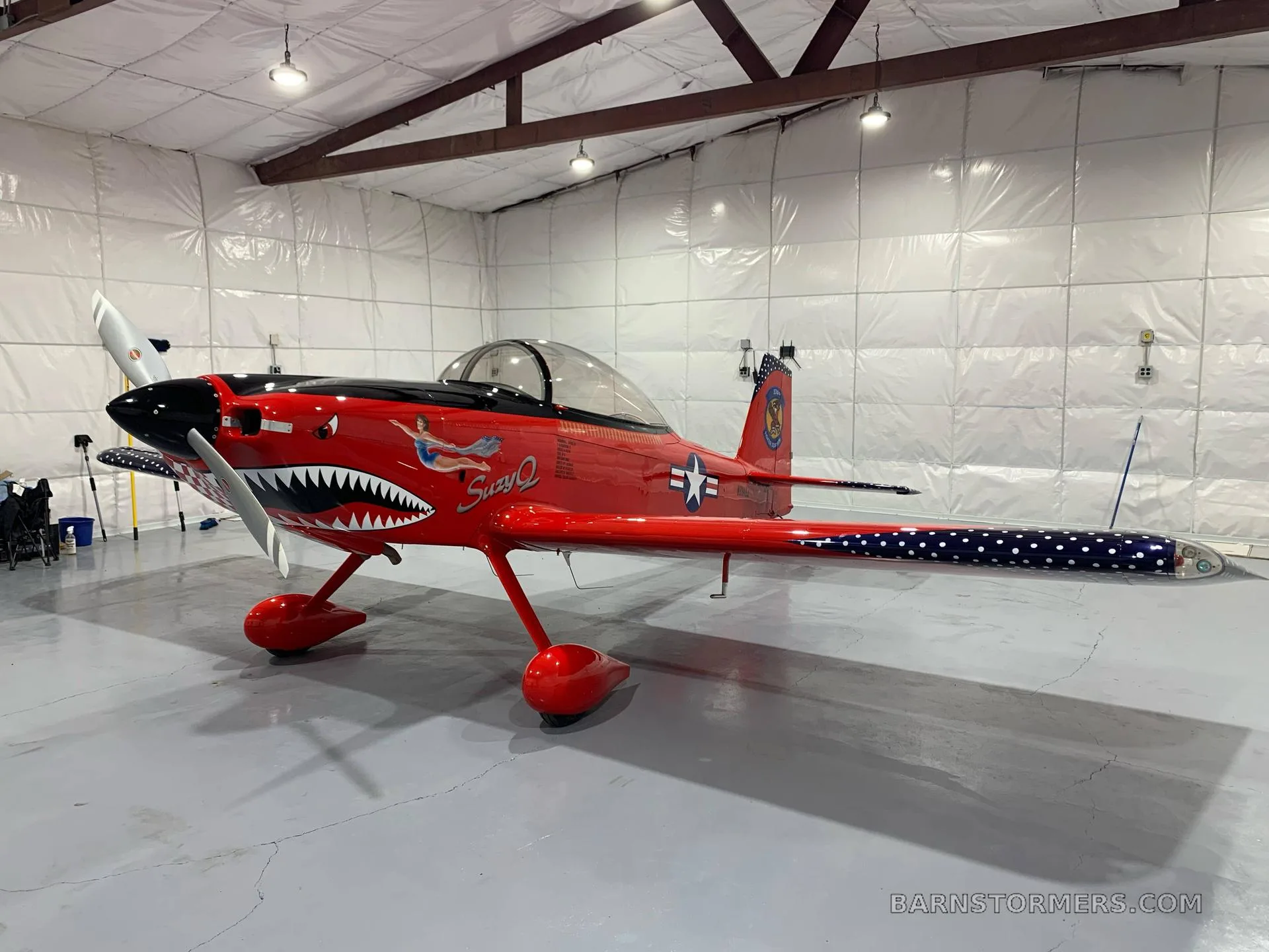 A red single-engine aircraft with a shark mouth nose art and patriotic wing tips, parked in a hangar.