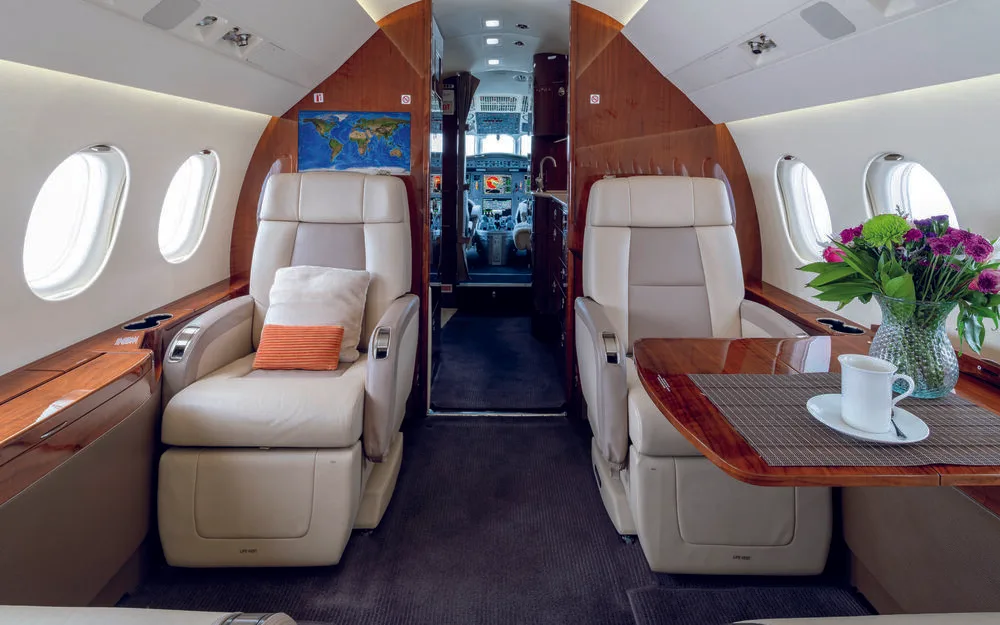 The spacious cabin interior of a business jet, with beige seats, a wooden table with flowers, and a view towards the cockpit through an open doorway.