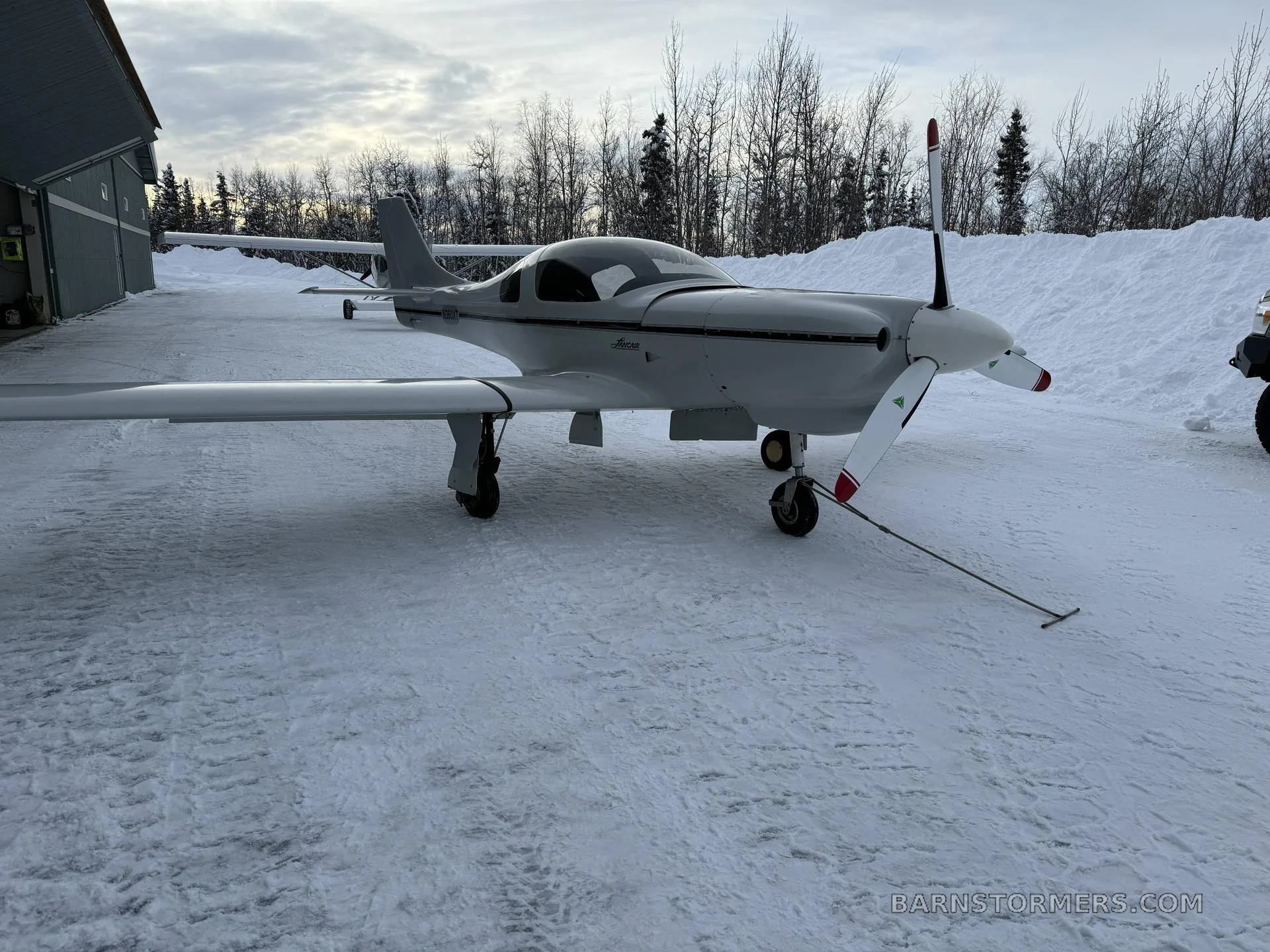 A white Lancair Legacy aircraft with a black stripe is parked on snowy ground next to a hangar, with trees in the background.