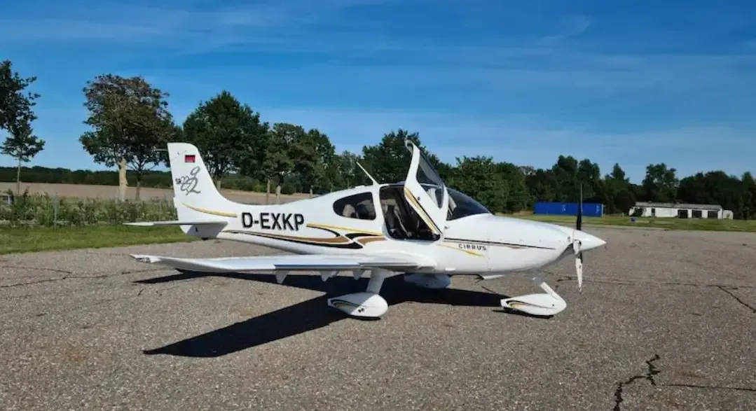A white Cirrus SR22 aircraft with gold and black stripes and registration D-EXKP parked on an asphalt surface with its doors open in a rural setting.