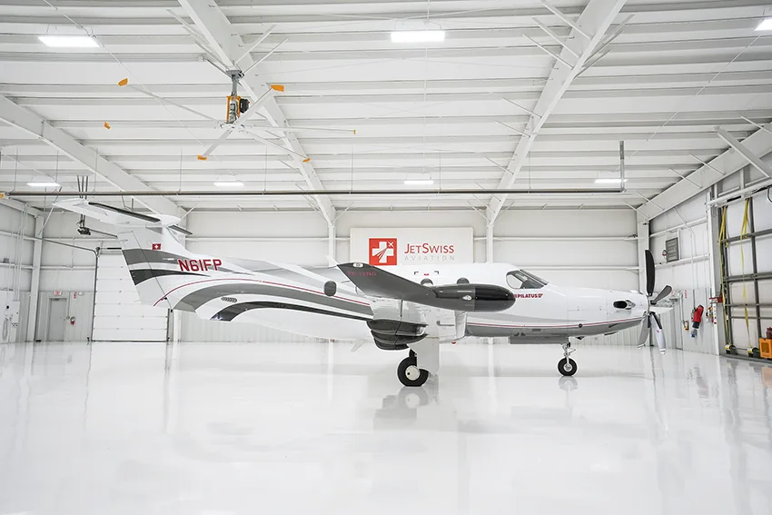 A white Pilatus PC-12 aircraft with gray and red stripes is seen from the side in a brightly lit hangar.