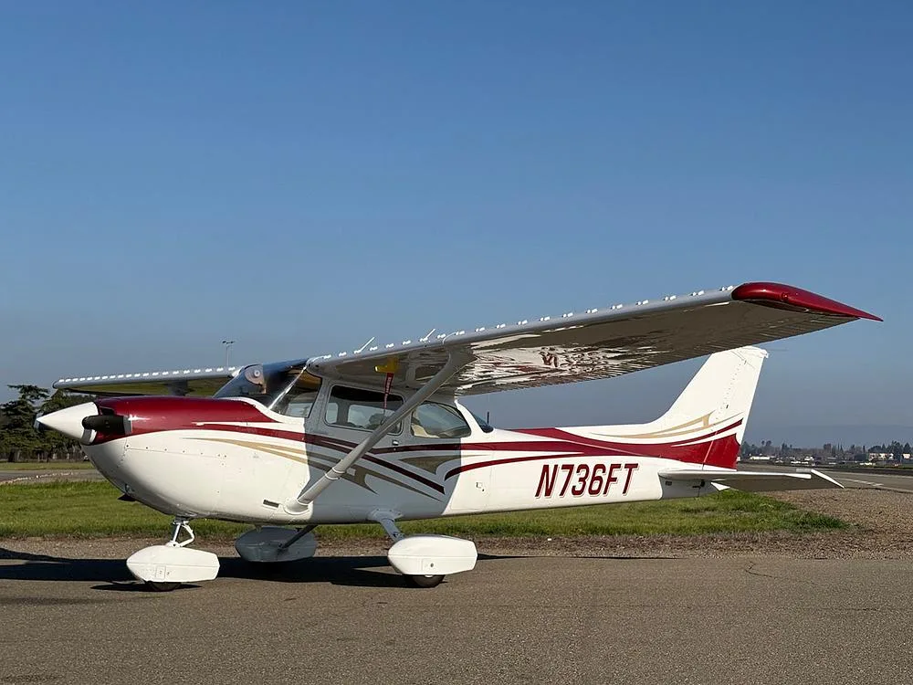 A white and maroon Cessna 172 with gold accents, parked on an airfield under a clear blue sky.