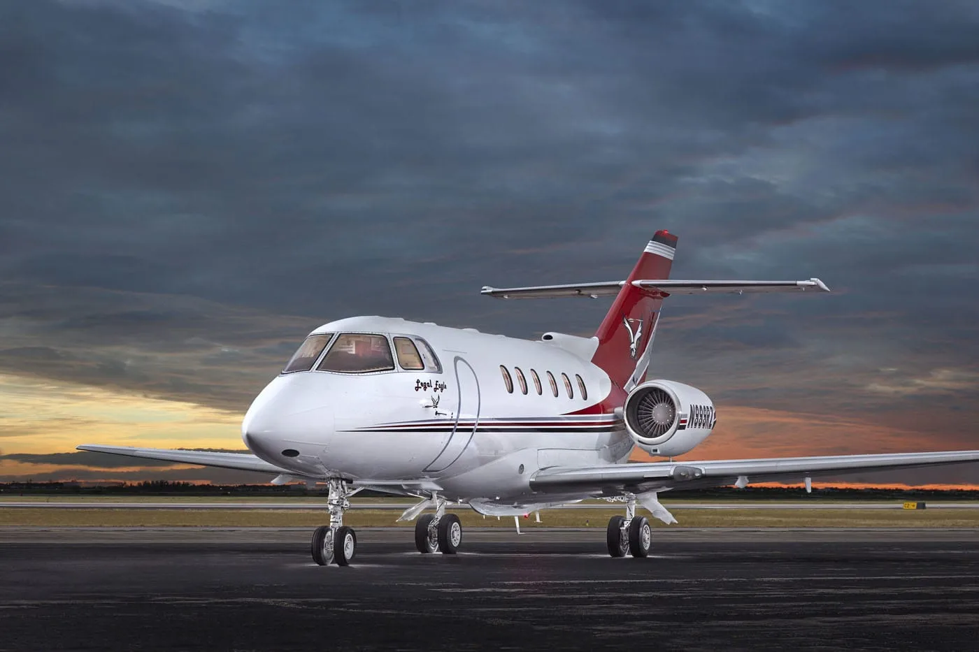 A white business jet with red and black accents on the tail and fuselage, parked on an airfield at sunset.