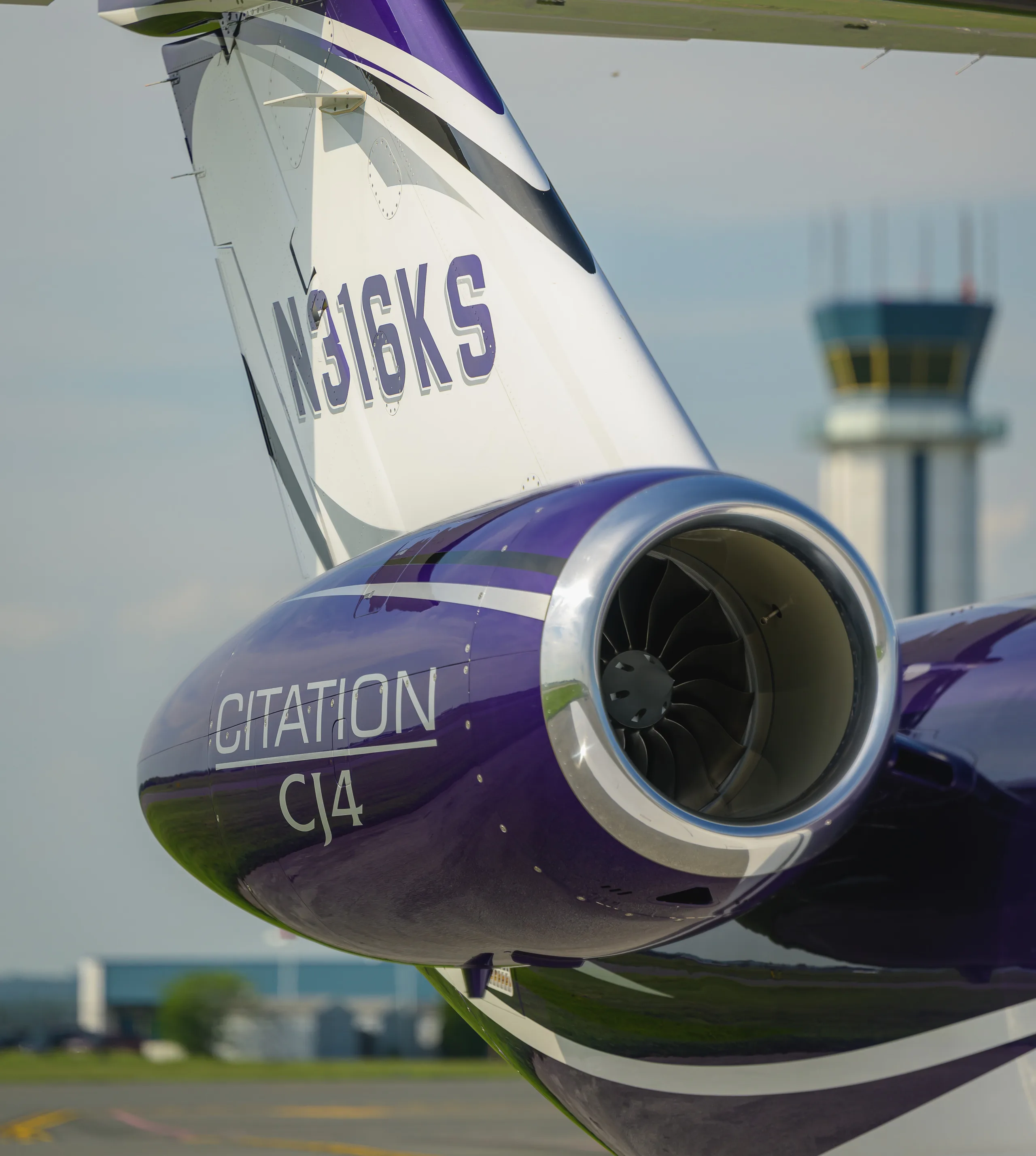 A close-up of the purple and white tail of a Cessna Citation CJ4 business jet, showing the N316KS registration and an engine nacelle, with an air traffic control tower in the background.