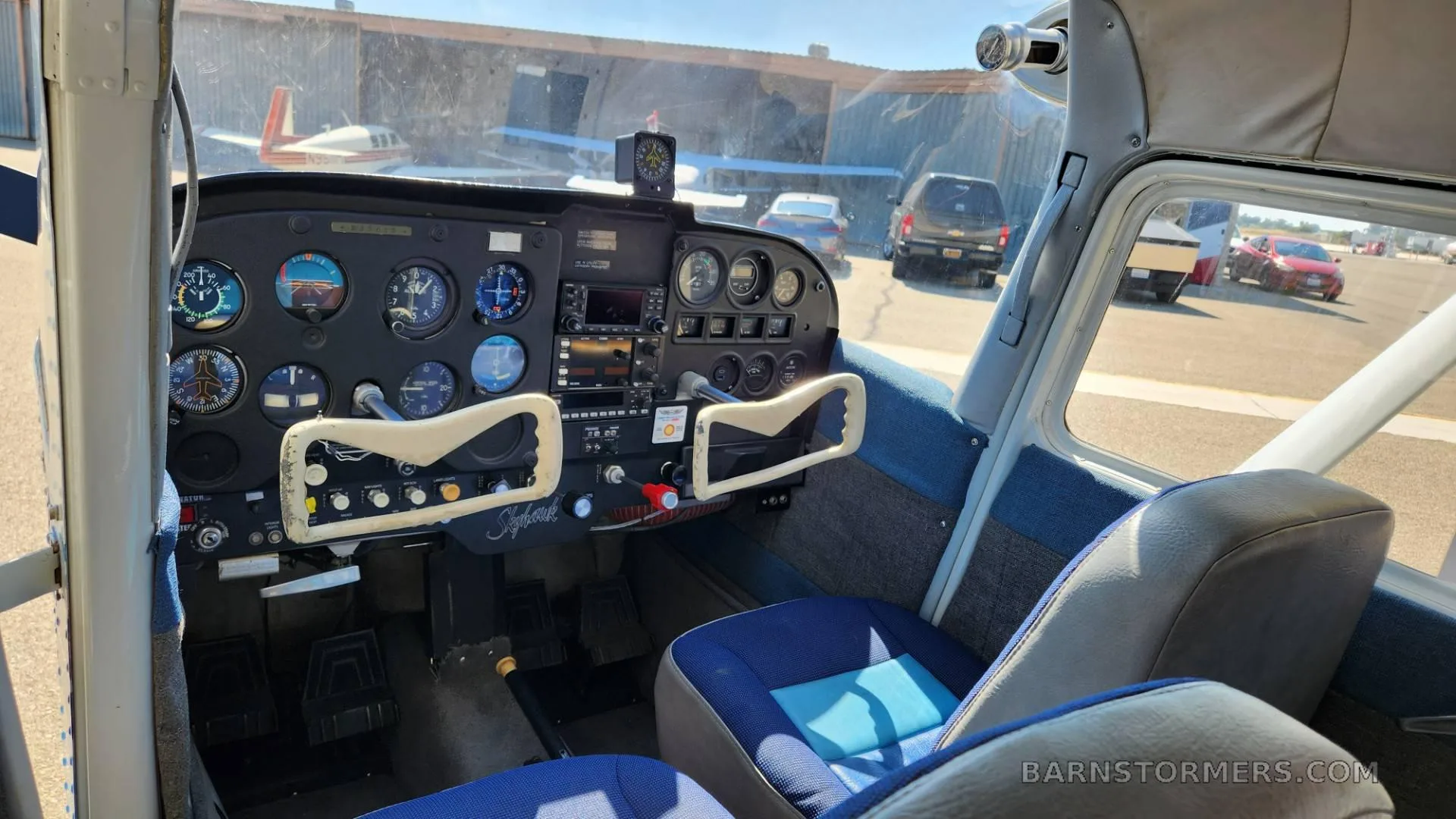 The cockpit of a light aircraft, showing the instrument panel, controls, and blue seats, with an airfield visible through the windshield.
