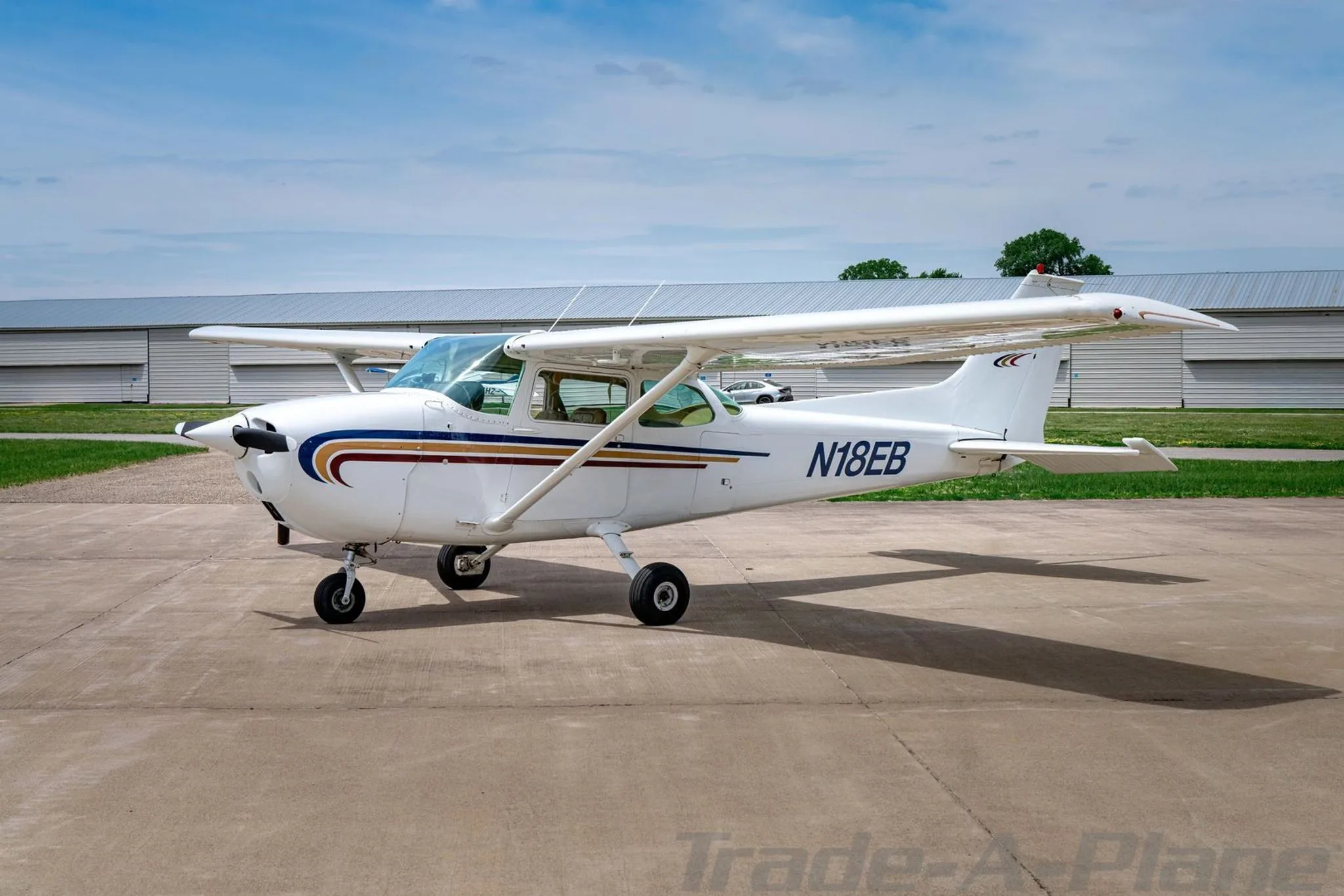 A white Cessna 172 with blue, gold, and red stripes on the nose and fuselage, parked on an asphalt ramp, viewed from the front-left.