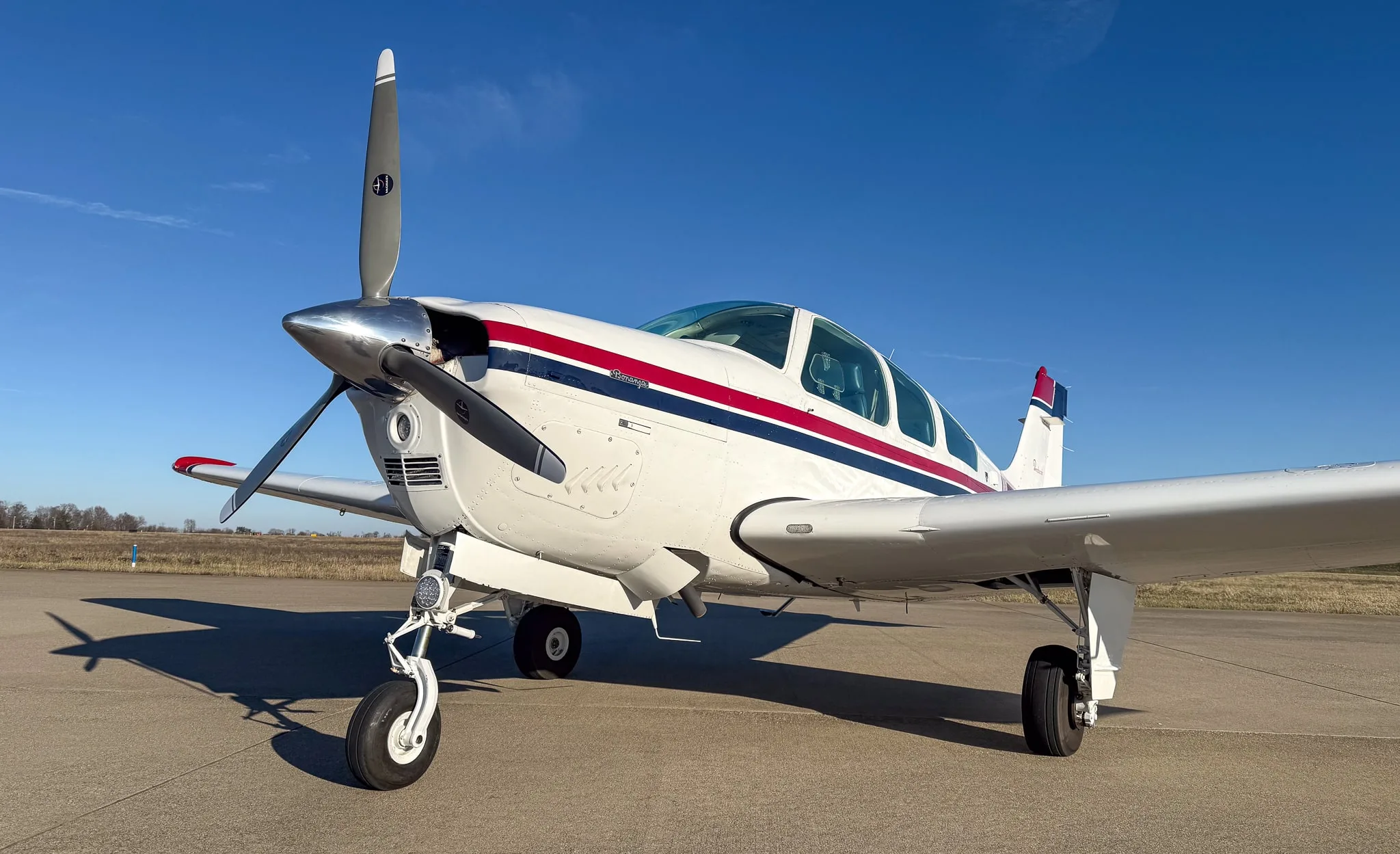 Low front-right view of a white Beechcraft Bonanza with red and blue stripes on an airfield.
