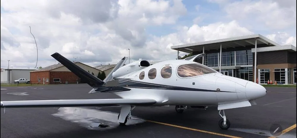 A white Cirrus Vision Jet with black and grey accents parked on an airport tarmac under a cloudy sky.