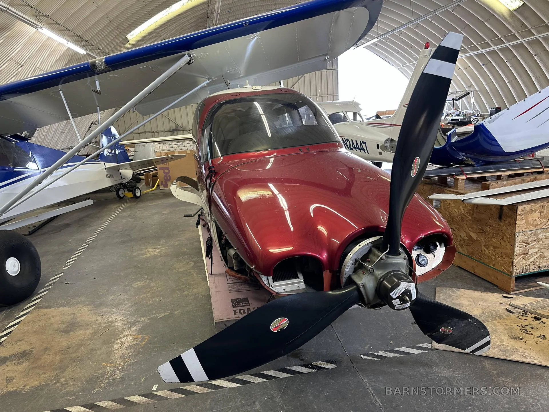 A red single-engine aircraft with its cowling removed, showing the propeller, inside a hangar with other aircraft.