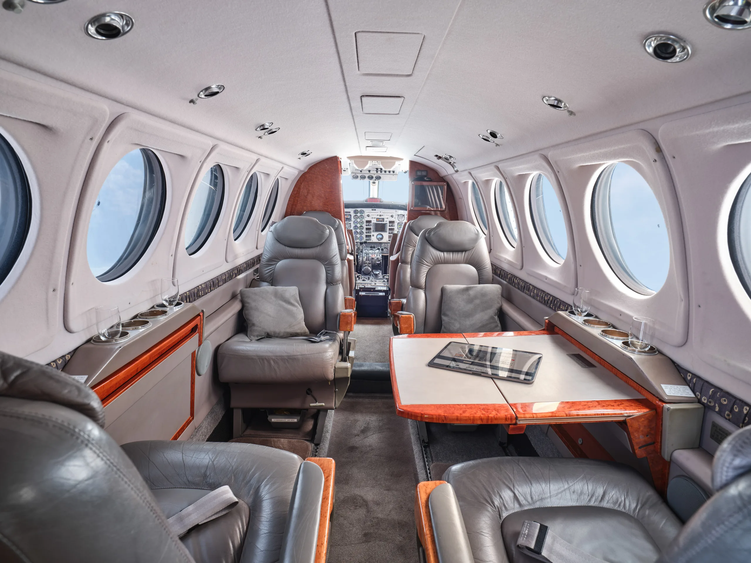 The luxurious cabin interior of an aircraft features grey leather seats, a wooden table, and a view into the cockpit.