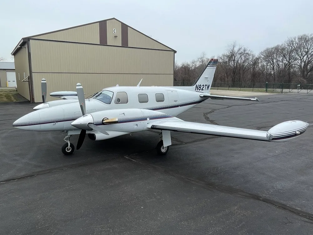 A white twin-engine aircraft, N82TW, with dark blue and red stripes, parked on a tarmac next to a hangar under an overcast sky.