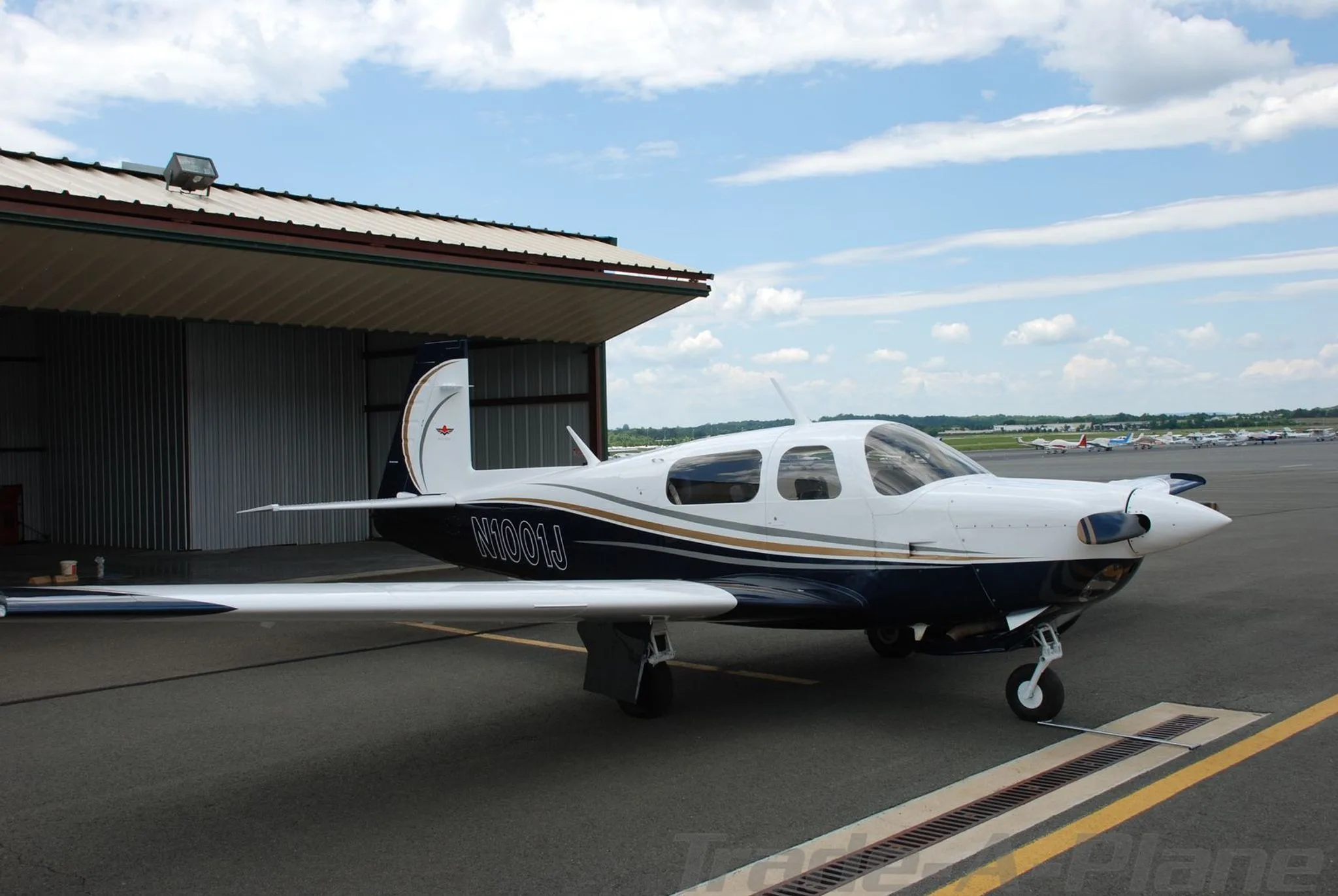 A white and blue Mooney aircraft (N1001J) parked on a tarmac next to an open hangar under a partly cloudy sky.