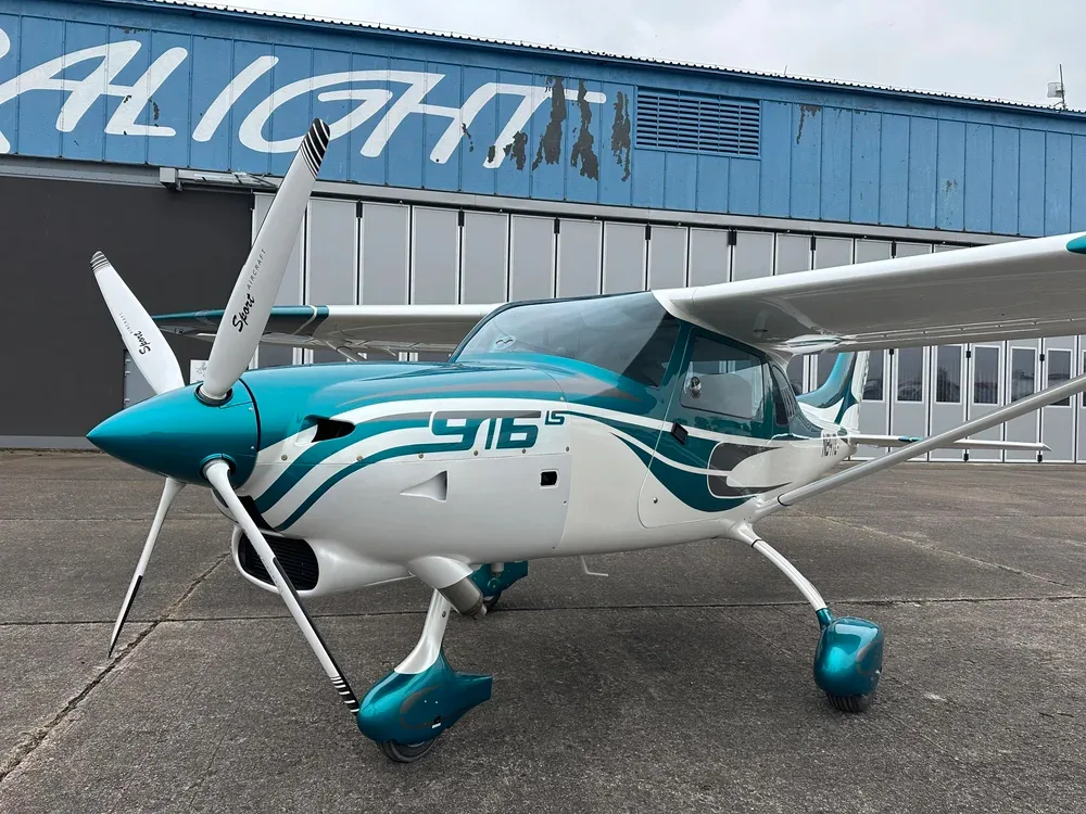 A close-up front view of a white and teal Sirius 916 LS aircraft, registration N84TL, parked in front of a blue hangar.