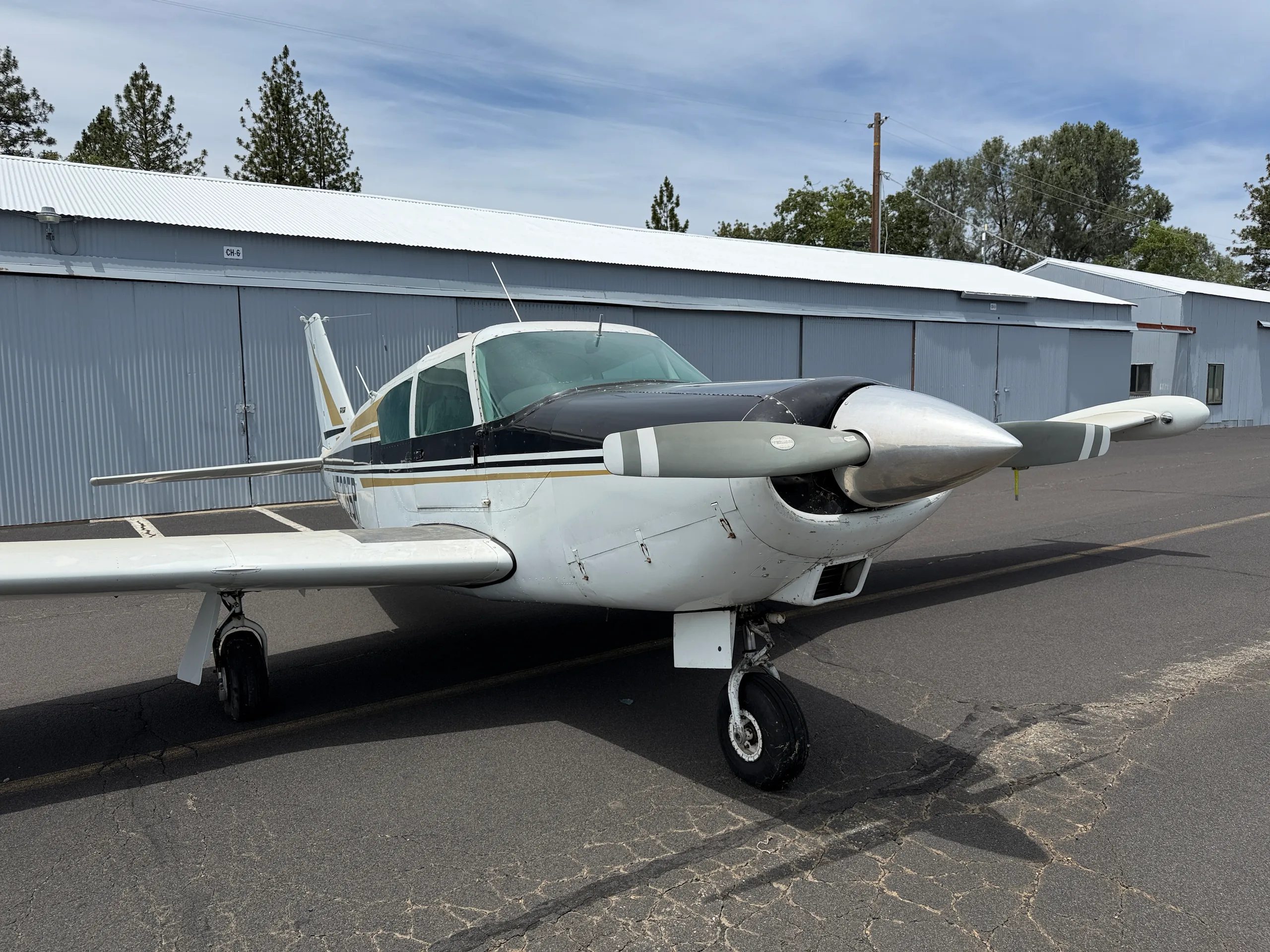 A white single-engine propeller aircraft with black and gold stripes is parked on an asphalt ramp in front of hangars.