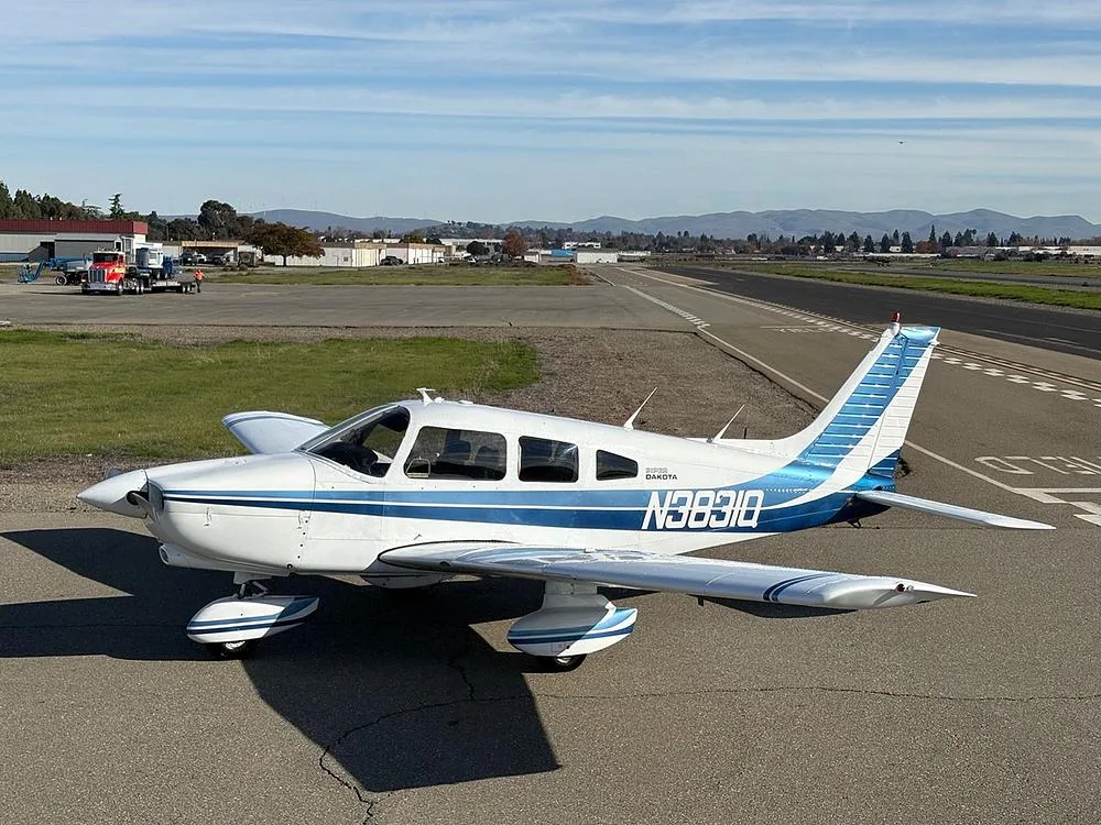 A white Piper Dakota aircraft with blue stripes, registration N3831Q, parked on an airfield with a runway and distant mountains.