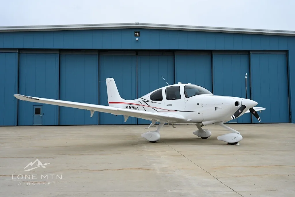 White Cirrus SR22 aircraft with red and black stripes, tail number N1071A, parked on an apron in front of a blue hangar.