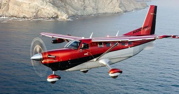 A red and white Quest Kodiak 900 aircraft flies low over blue water with rocky cliffs in the background.