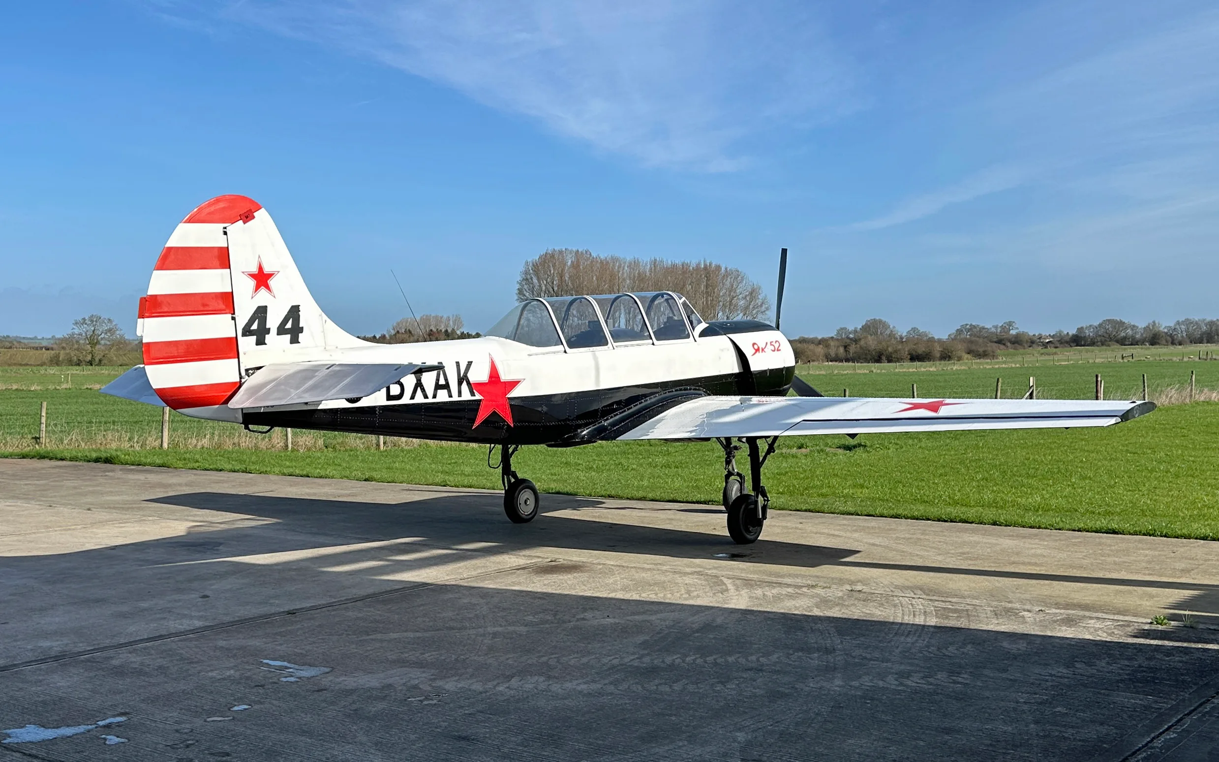 A white and black Yak-52 aircraft with red stars and tail stripes, parked on a concrete pad next to a grassy field.