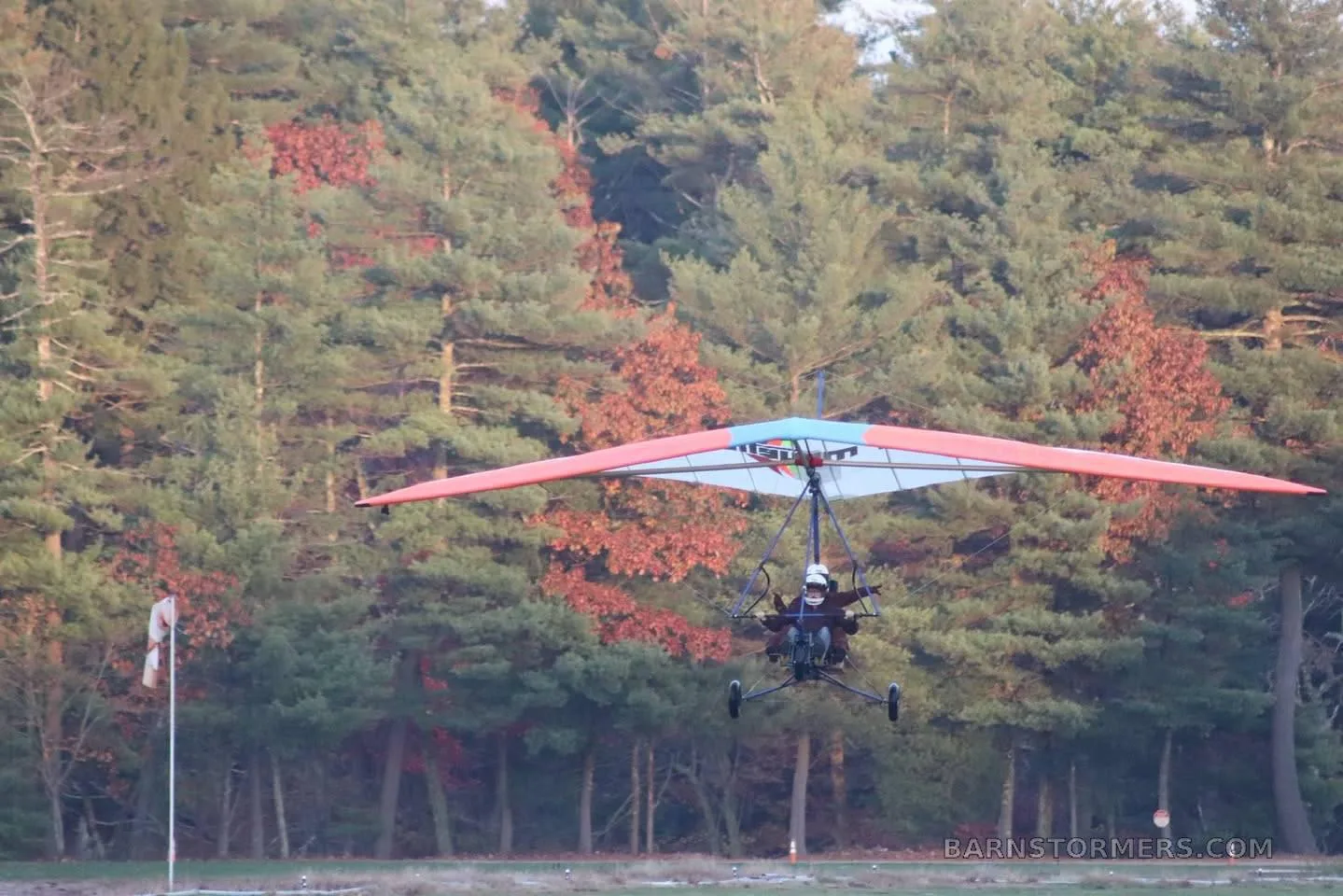 A powered hang glider with an orange, blue, and white wing flies low over a field with a dense forest in the background.